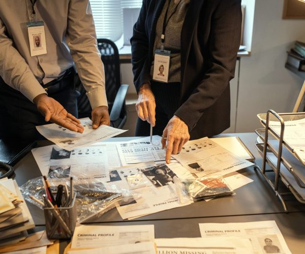Hands of intercultural agents of FBI discussing criminal profiles by workplace Hands of intercultural agents of FBI discussing criminal profiles by workplace while one of them pointing at photo of suspect